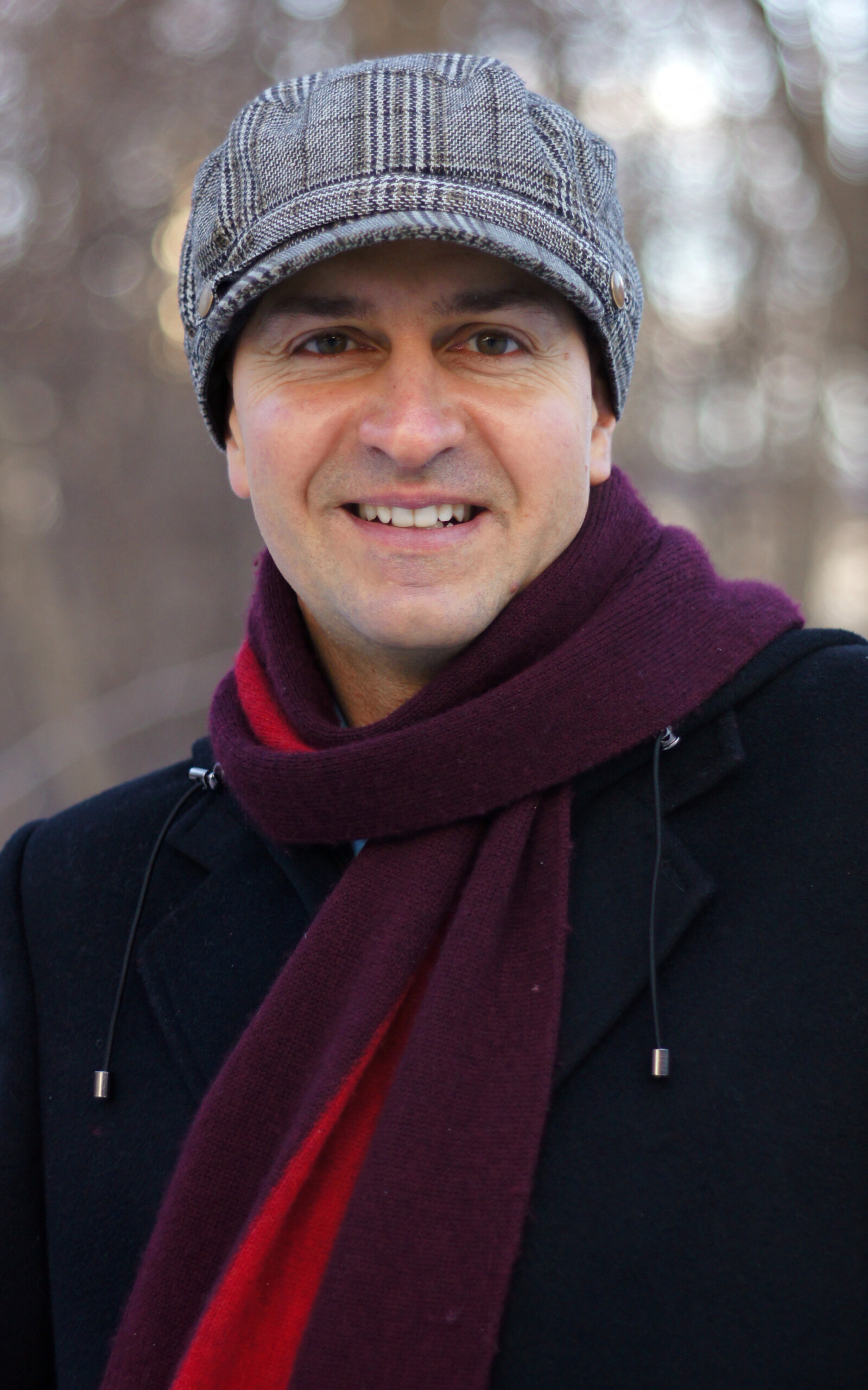Portrait of Sudarsana (Spiros Tsiaousis) smiling outdoors, wearing a grey cap and maroon scarf.
